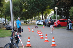 Pylone auf der Strecke in der Mitte des Bildes am Rande PKW-Parkplätze drei Leute mit Rückenansicht beobachten ein Kind auf dem Laufrad beim Parcour.