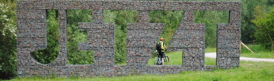 Im Grünen ein Bausteinkunstwerk mit ausgestanzten Buchstaben; JETZT. im Z ist ein Mountainbiker zu sehen